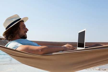Smiling man using laptop while relaxing on hammock in beachの写真素材