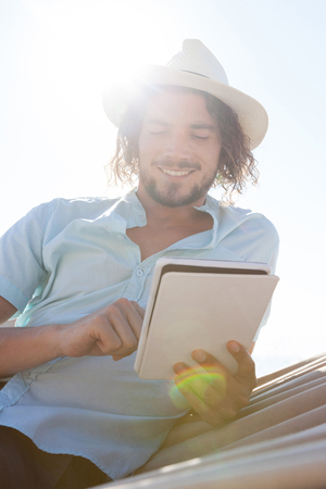 Smiling man relaxing on hammock and using digital tablet on the beachの写真素材