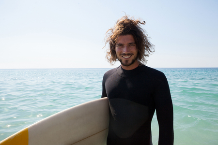 Portrait of smiling surfer with surfboard standing at beach coastの写真素材
