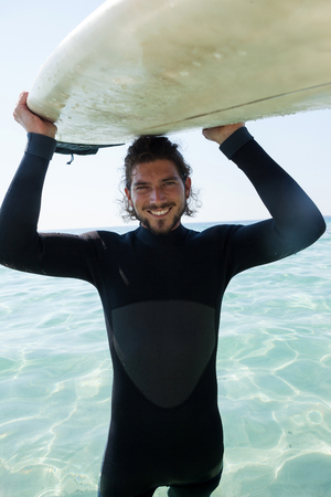 Portrait of smiling surfer in wetsuit carrying surfboard over head at beach coastの写真素材