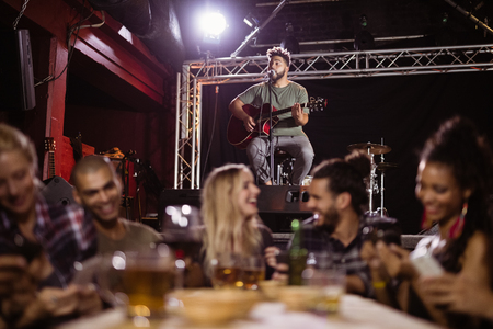 Smiling friends sitting by table with musician in background at nightclubの写真素材