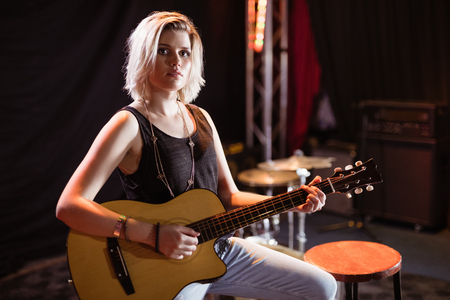 Portrait of female musician playing guitar while sitting on stool in nightclubの写真素材