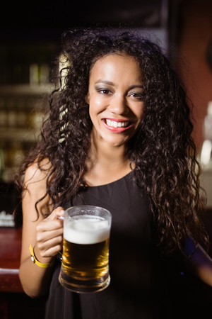 Portrait of smiling woman with beer glass standing by counter in nightclubの写真素材