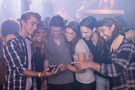 Smiling friends looking at smart phone while standing in nightclubの写真素材