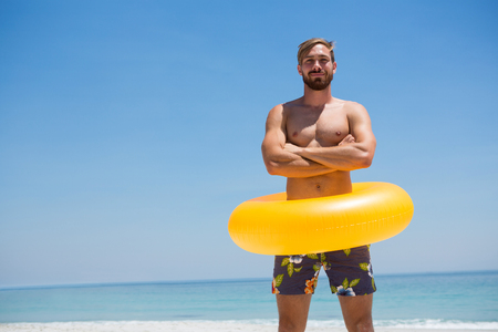 Portrait of man with arms crossed wearing inflatable ring while standing at beach on sunny dayの写真素材