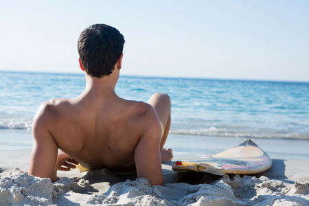 Rear view of shirtless man reclining by surfboard on sand at beach during sunny dayの写真素材