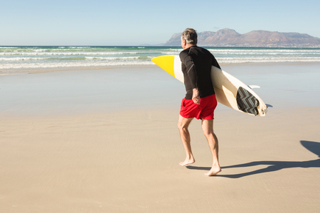 Rear view of man carrying surfboard while running on sand at beachの写真素材