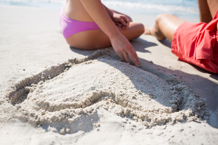 Mid section of young couple sitting by heart shape carved on sand at beach during sunny dayの写真素材