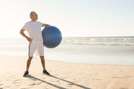 Senior man holding exercise ball while standing at beach on sunny dayの写真素材
