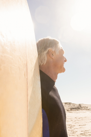 Close up of senior man with surfboard standing against sky at beachの写真素材