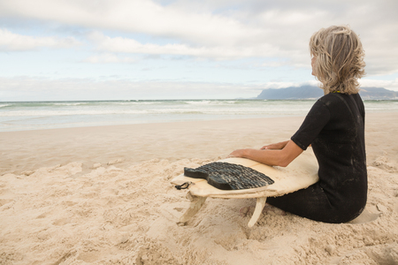 Woman in wetsuit sitting with surfboard against cloudy sky at beachの写真素材