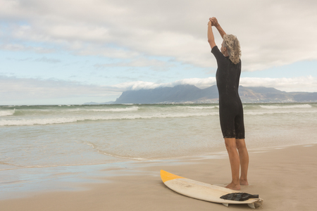Rear view of woman stretching while standing by surfboard against cloudy sky at beachの写真素材