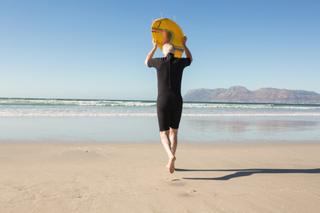 Rear view of senior man carrying surfboard at beach on sunny day