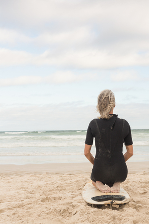 Rear view of woman kneeling on surfboard against sea at beachの写真素材
