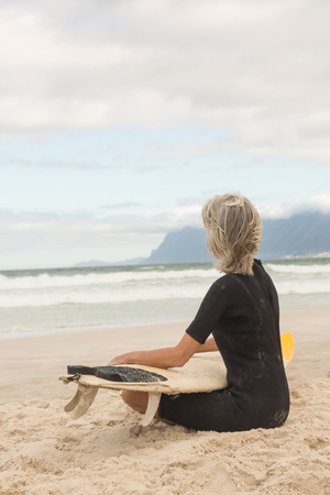 Side view of woman in wetsuit sitting with surfboard against cloudy sky at beachの写真素材