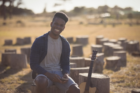 Portrait of man holding smart phone while sitting on tree stump on sunny dayの写真素材