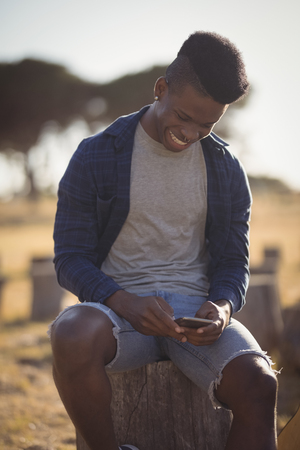 Smiling man using smart phone while sitting on tree stump against skyの写真素材