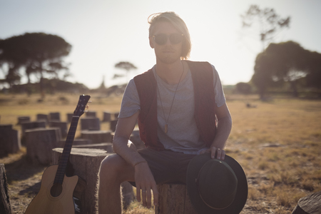 Portrait of young man sitting by guitar on tree stump against clear skyの写真素材