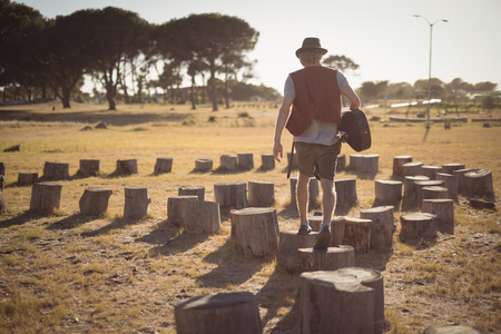 Rear view of man holding guitar while walking on tree stumps at fieldの写真素材