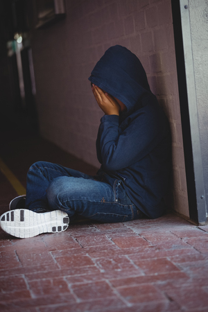Sad boy sitting on pavement by wall in corridor at schoolの写真素材