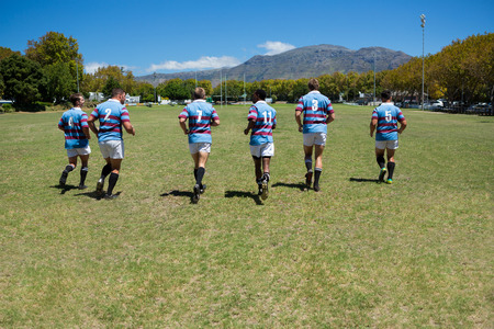 Rear view of rugby team running at field on sunny dayの写真素材