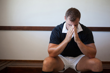 Thoughtful rugby player sitting on bench against white wall in locker roomの写真素材