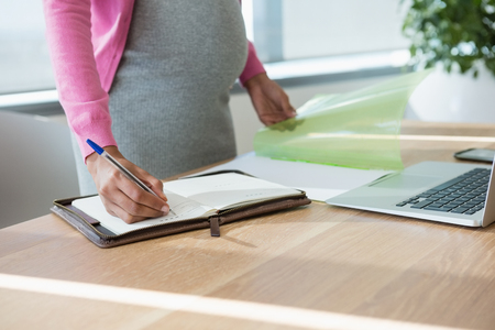 Mid section of pregnant businesswoman writing in organizer at desk in officeの写真素材