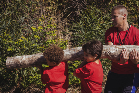 Trainer assisting kids in carrying a heavy wooden log during obstacle course training at boot campの写真素材