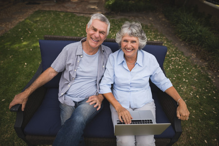 High angle portrait of smiling senior couple sitting together with laptop on couch in backyardの写真素材