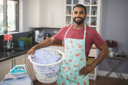 Portrait of smiling young man holding laundry basket by ironing board in kitchen at homeの写真素材