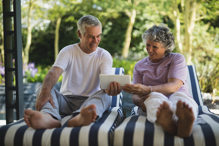 Happy senior couple using digital tablet while sitting on lounge chairs