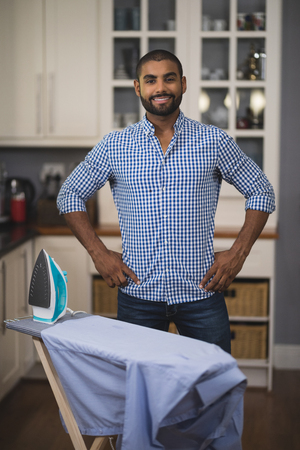 Portrait of smiling young man standing by iron on board in kitchen at homeの写真素材
