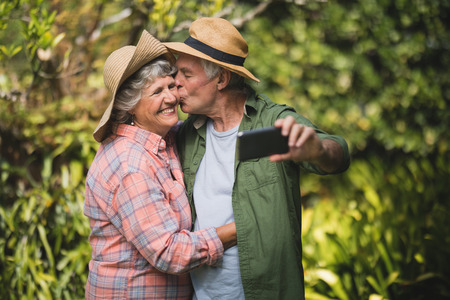 Senior man kissing smiling woman while taking selfie against plants