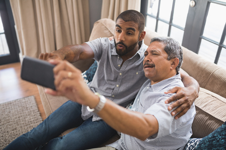 Happy man with his father taking selfie while sitting together on couch at homeの写真素材