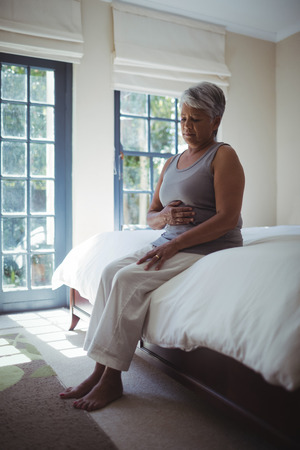 Upset senior woman touching belly on bed at homeの写真素材