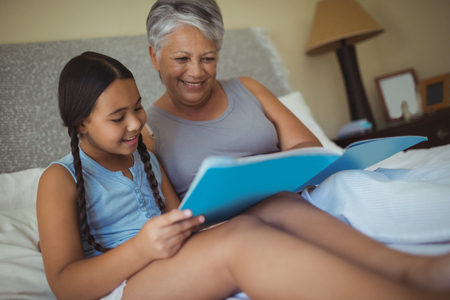 Grandmother and granddaughter watching photo album together in bed room at homeの写真素材