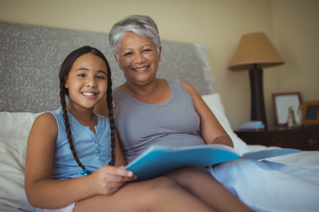 Portrait of grandmother and granddaughter holding photo album in bed room at homeの写真素材