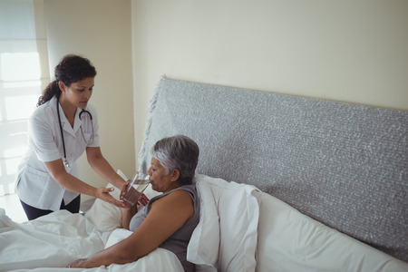 Female doctor giving sick senior woman a glass of water on bed at home