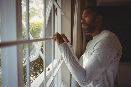 Side view of thoughtful man looking out through window while standing at homeの写真素材