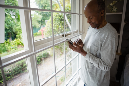 Senior man using mobile phone while standing by window at homeの写真素材