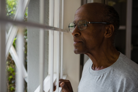 Thoughtful senior man wearing eyeglasses while looking out through window at home