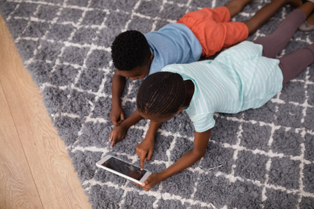 High angle view of siblings using digital tablet while lying on rug at homeの写真素材