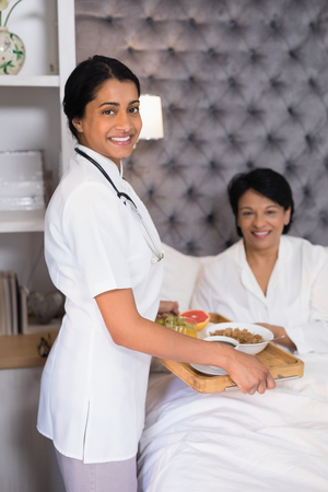 Portrait of smiling nurse giving breakfast to patient resting on bed at homeの写真素材