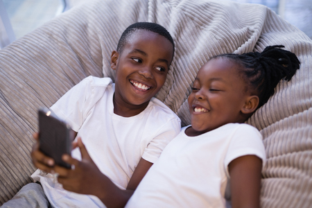 High angle view of smiling siblings using mobile phone while lying on couch at homeの写真素材