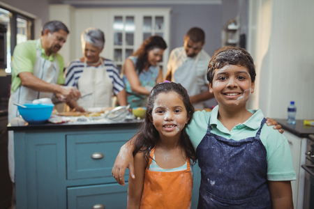 Siblings smiling at camera while family members preparing dessert in background at homeの写真素材