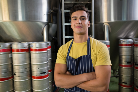 Portrait of smiling male worker with arms crossed standing by storage tanks at breweryの写真素材