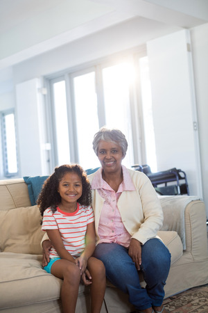 Smiling grandmother and granddaughter sitting on sofa in living room at homeの写真素材