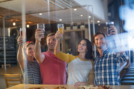 Cheerful friends with hand raised holding beer glass while standing at table in barの写真素材