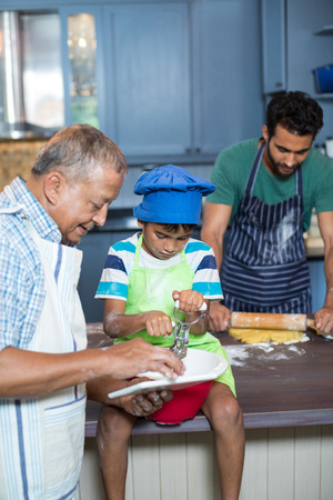 Grandfather showing tablet computer to boy with man rolling dough in background