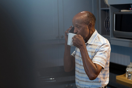 Man having coffee while talking on phone in kitchen at homeの写真素材